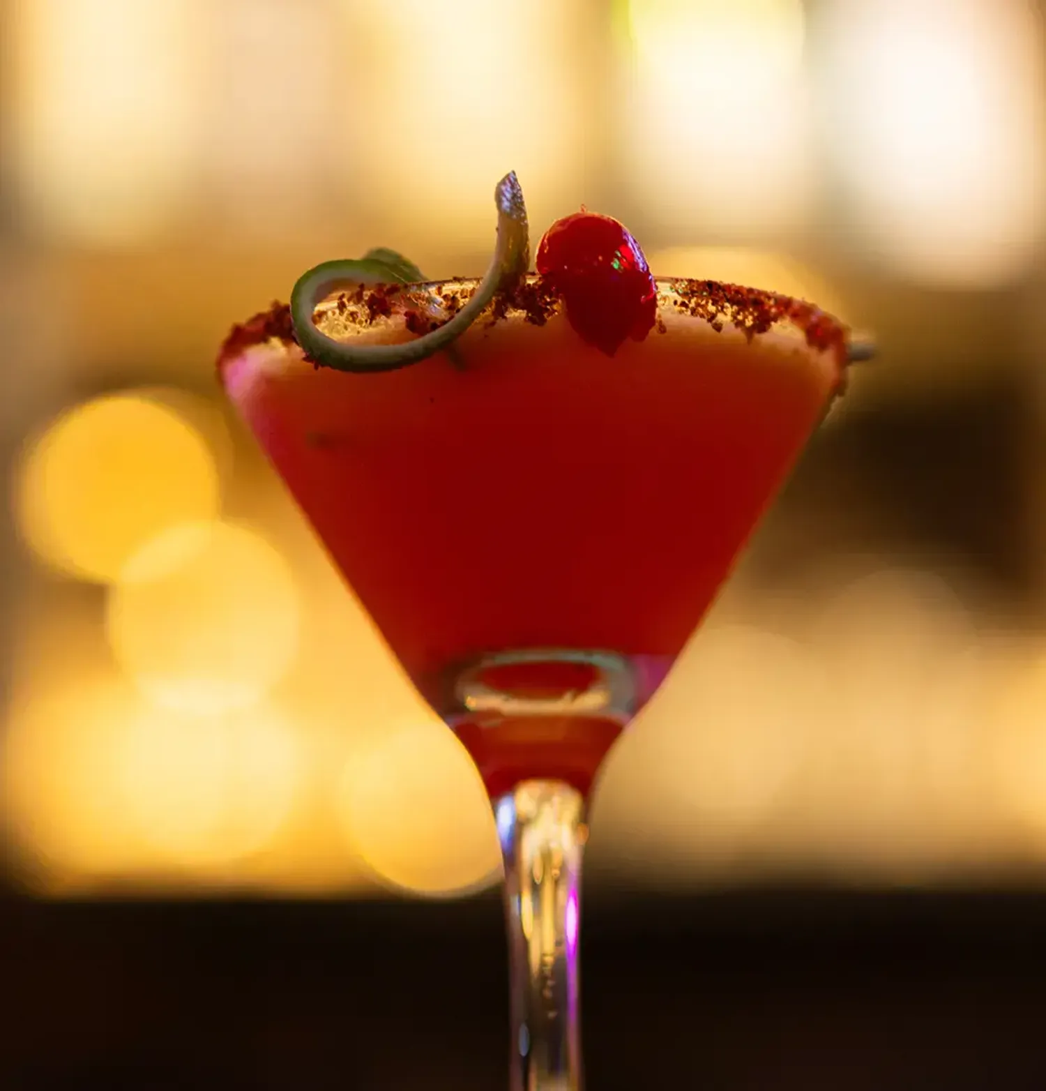 Red cocktail in a martini glass with chili frosting, cherry, and lemon peel garnish