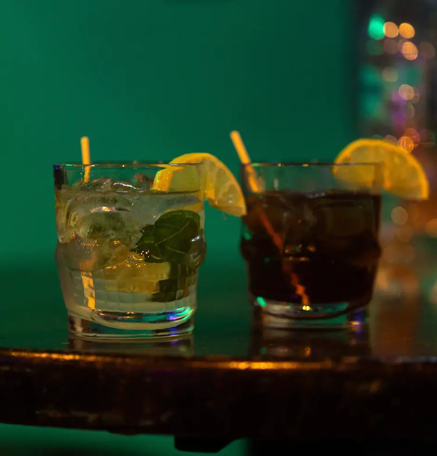 Two cocktails in a short glass, one light with mint and one dark, both garnished with a lemon slice