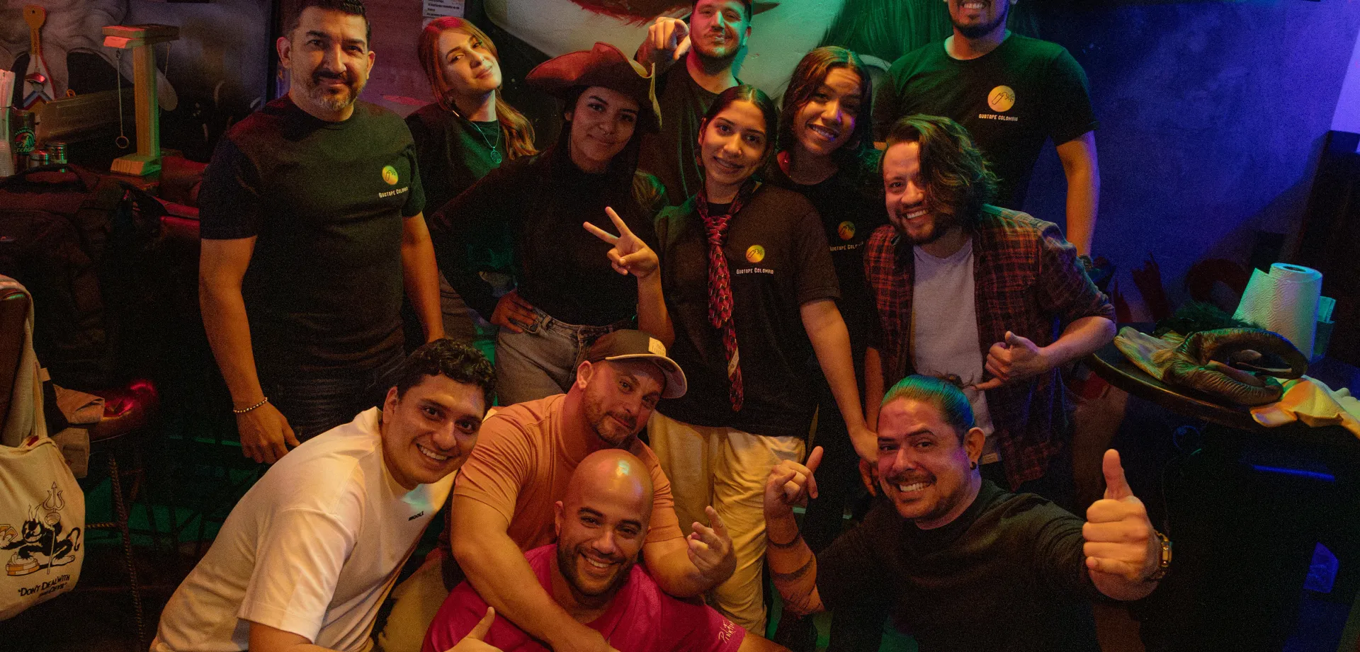 Group of friends posing together and smiling in a dimly lit bar with colorful green and red lights, standing in front of a large mural of a clown face.