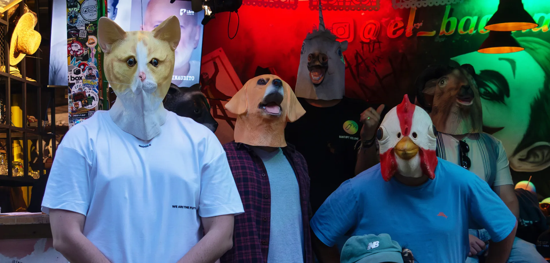 Group of people wearing realistic animal masks (cat, dog, horse, rooster, and monkey) posing together inside a neon-lit bar with hanging lamps and colorful decorations.