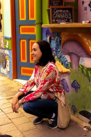 A woman crouches in front of a colorful mural next to a sign advertising "Cerveza Cannabis Weed Beer."