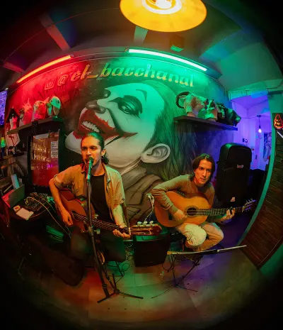 Two musicians playing acoustic guitars in front of a large Joker mural and a neon sign "@el_bacchanal" in a venue with red and green lights.