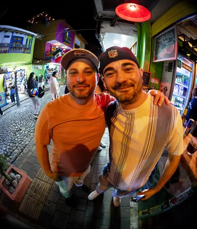 Two smiling, bearded men pose embracing on a cobblestone street with brightly colored colonial buildings in the background.