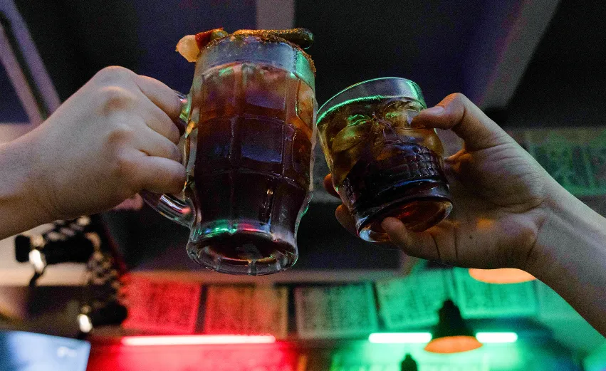 Two people clinking a cocktail mug and a skull-shaped glass with drinks in a festive, neon-lit bar.
